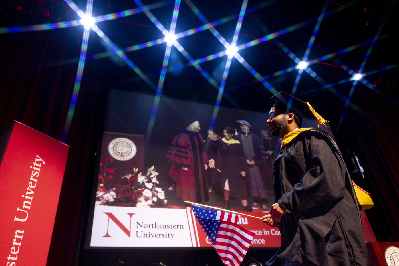 A graduate in a cap and gown holds in an American flag while crossing the stage at a commencement ceremony. 