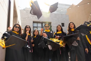 A group of people in graduation attire smile and toss their caps into the air while standing outdoors in bright sunlight.
