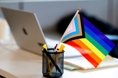 A Progress Pride flag displayed on an office desk next to a MacBook.