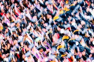 A large crowd of people in graduation gowns and caps is captured in colorful, energetic motion during a ceremony.