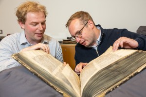 Two historians examine a large medieval manuscript inside a library.