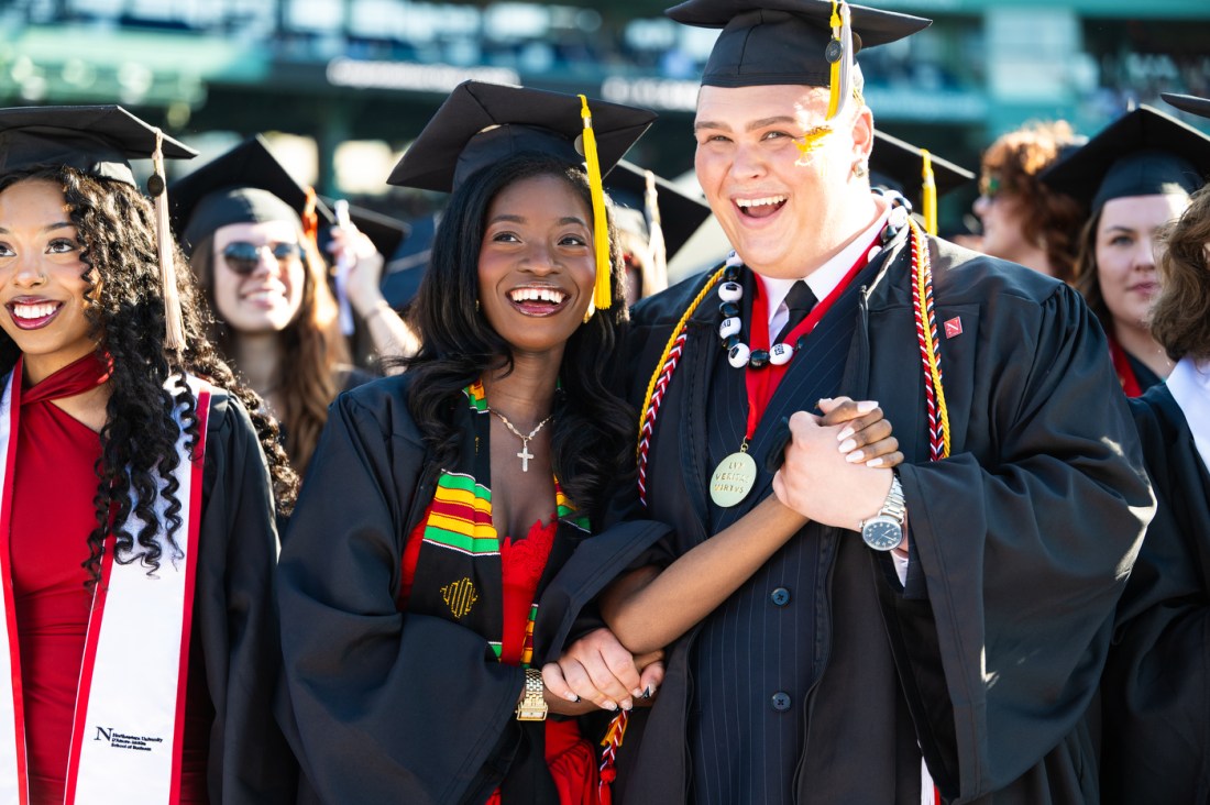 A group of jovial Northeastern University graduates in cap and gowns stand together on the baseball diamond at Fenway Park.