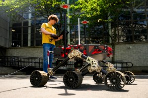 A person stands outdoors working on a large wheeled robotic device, with a laptop in hand and trees and a building in the background.