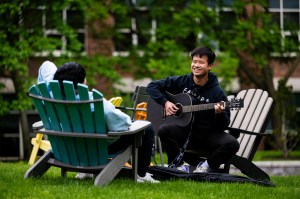Two students sit in Adirondack-style chairs on Centennial Common on the Boston Campus. One of them plays guitar.