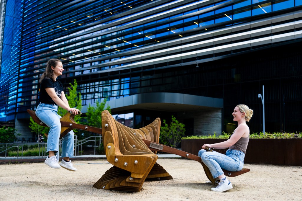 Two people enjoy a moment on a wooden seesaw in an outdoor space, with a modern building and greenery in the background.
