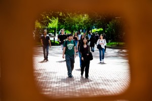 People walk across a sunlit brick pathway surrounded by trees and shadows.