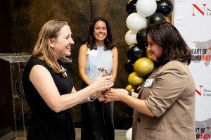 Two people shake hands in a congratulatory moment at the Heart of Community Awards ceremony. A third person can be seen smiling in the background standing next to black, white, and gold balloons.