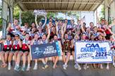 Members of the womens row team sit on a stage cheering after winning the CAA finals. They are holding signs that say &#039;CAA Champions 2025&#039; and &#039;CAA Ticket Punched&#039; on them.