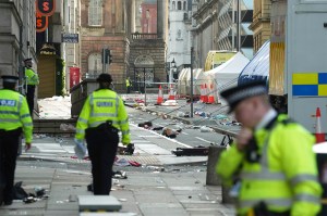 Police officers walking around a road examining debris after a car collided with pedestrians at a parade.