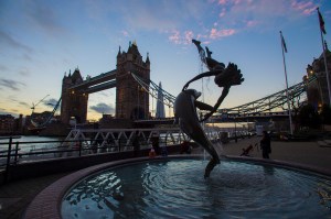 A sculpture of a girl with a dolphin in front of the Tower Bridge at sunset.