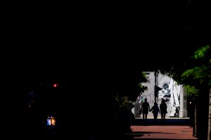 Silhouetted figures walk toward a brightly lit space framed by shadows and surrounding structures.