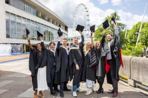 Seven graduates smiling while throwing their caps in the air. The London Eye ferris wheel is visible in the background. It is a bright sunny day.
