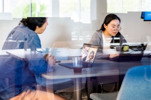 Two people collaborate at a table while others appear on a laptop screen nearby.