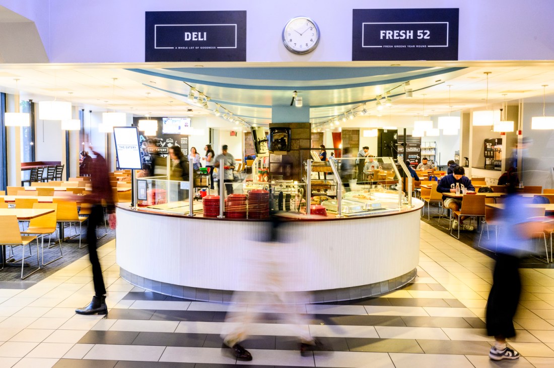 Students walk through a brightly lit Northeastern campus dining hall featuring deli and salad stations. 