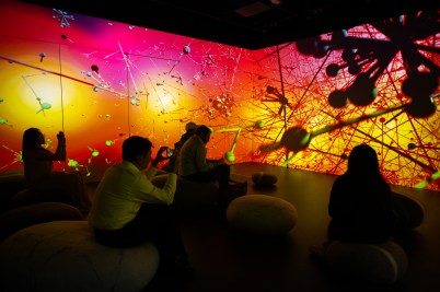 People sit in a dark room surrounded by colorful, abstract visuals projected on the walls.