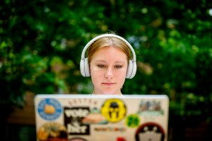 A person wearing headphones looks at a laptop covered in colorful stickers, with greenery in the background.