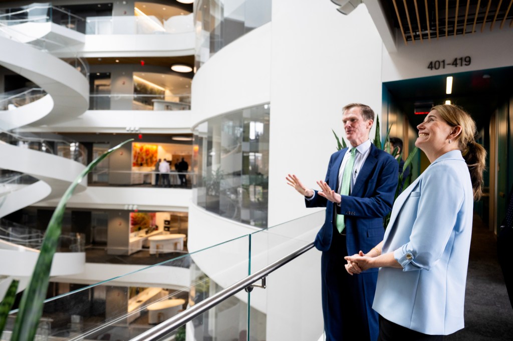 Two people stand indoors near a modern glass railing, looking out across a large, multi-level building interior.