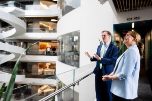 Two people stand indoors near a modern glass railing, looking out across a large, multi-level building interior.