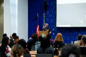 A view of an indoor event with an audience listening to a speaker at a podium, with flags and a large screen in the background.