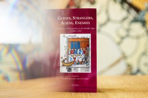 A copy of the book “Guests, Strangers, Aliens, Enemies: Ambiguities of Hospitality in the Middle Ages, c. 1000–1350” is displayed on a table with a decorative background.