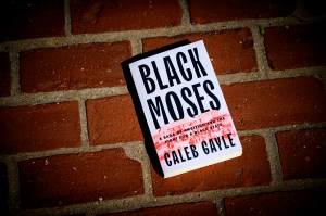 A copy of 'Black Moses' by Caleb Gayle lying flat on a red brick surface. The book cover features bold black text on a white background. The book appears to be hardcover with a dust jacket.