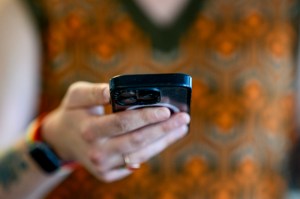A person holds a smartphone, with the screen out of view and a patterned orange and green shirt in the background.