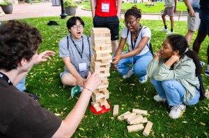 Four people sit on grass around a tower of wooden blocks, reacting to a tense moment during a game outdoors.