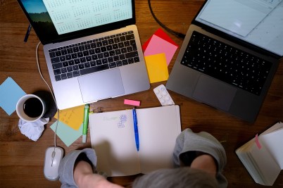 Person sitting at a cluttered desk with two open laptops, colorful sticky notes, a notebook, coffee, and scattered office supplies.