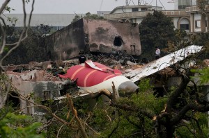 Wreckage of a commercial plane lies on top of a damaged building surrounded by debris and broken trees.