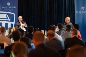 President Aoun sitting on stage next to another speaker in front of a crowd of audience members at the AI Synergy Summit.