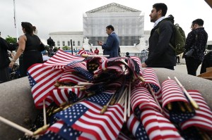 People gather outside the U.S. Supreme Court, with a pile of small American flags in the foreground and the courthouse under partial scaffolding in the background.