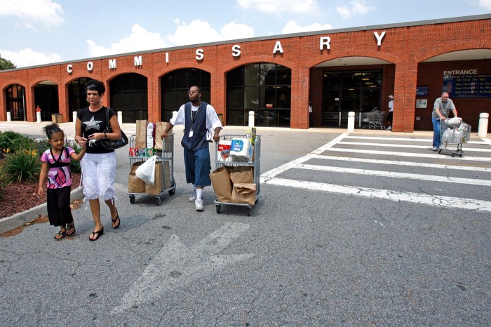 A group of people walking, some moving mobile carts, outside a brick building with "COMMISSARY" spelled out in large white letters across the top