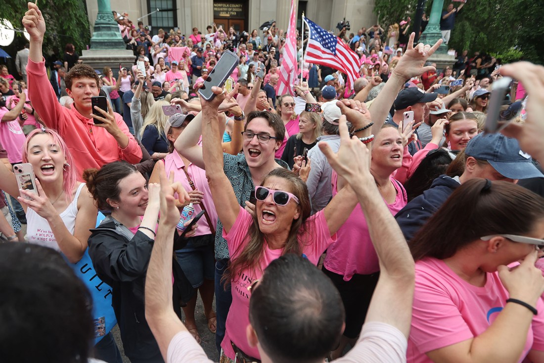A crowd of supporters, many wearing pink articles of clothing or accessories, cheering with their arms raised over their heads in celebration of the Karen Read trial verdict.