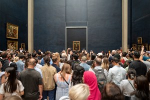 A large crowd gathers in front of the Mona Lisa at the Louvre Museum, with many visitors holding up phones to take photos.