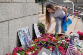 A person places flowers at a growing memorial site on the steps of a public building, surrounded by colorful bouquets, American flags and tribute signs.
