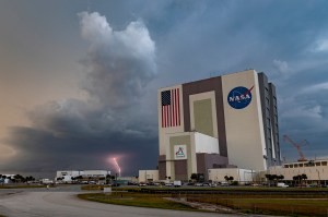 NASA's Vehicle Assembly Building at the Kennedy Space Center with an American flag and NASA logo visible on the exterior, set against a dramatic cloudy sky.
