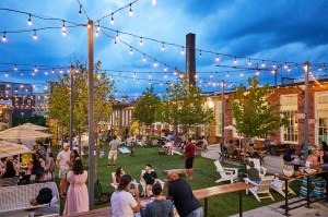 An outdoor evening scene showing a bustling public plaza with string lights overhead and landscaped areas with trees and greeny, scattered seating, and people gathered socializing.