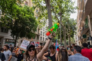 A person wearing a baseball cap and sunglasses holds a watergun up in the air with one hand. They are standing amongst a large crowd of people protesting against tourism.