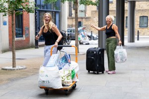 Two people smile while unloading bags and luggage outside a building on a city sidewalk.