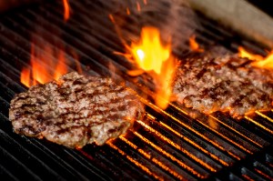 Two burgers on a grill, flames leaping up around them. They have char marks on them.