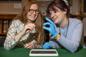 Sue Jones and Rebecca Raven sitting at a table looking at an old coin. The person holding the coin is wearing blue gloves.