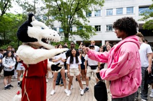 A person in a costume interacts with a student in a crowd outside on a campus walkway.