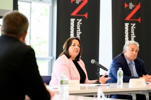 A person speaks at a table during a formal discussion event, with others seated nearby.