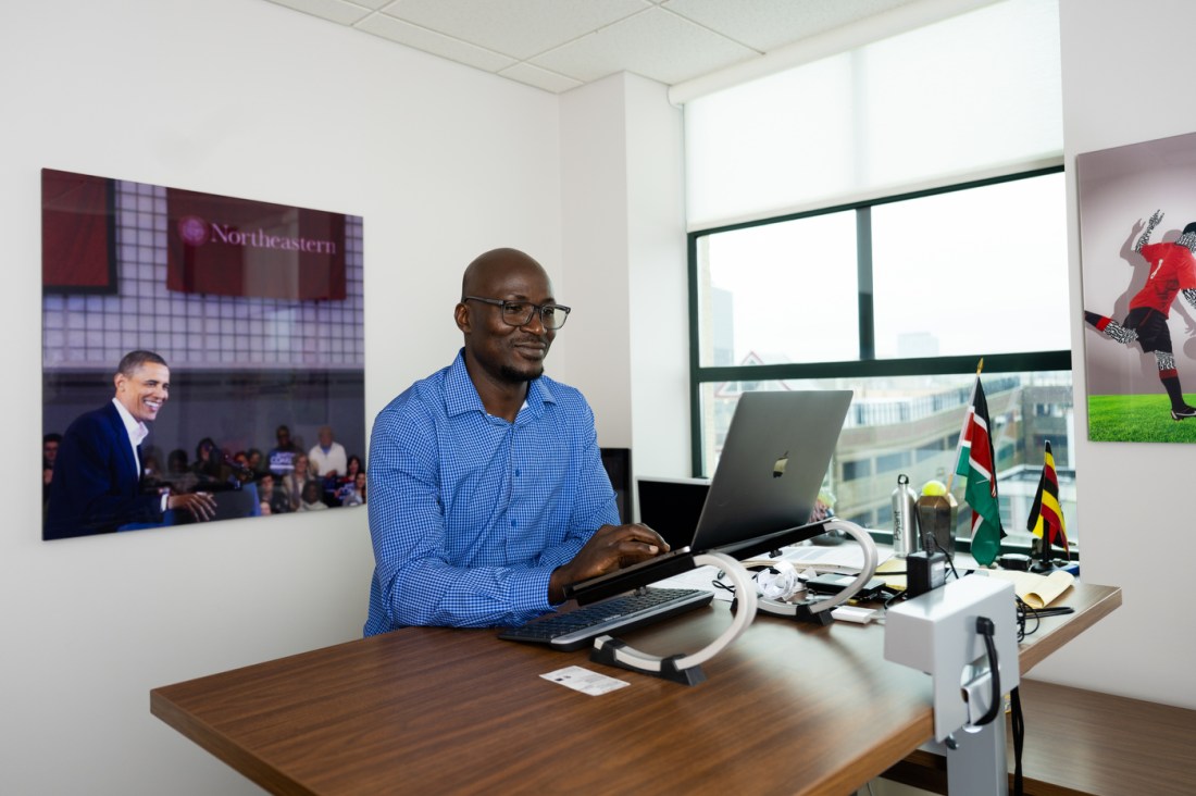Francis Oywech sitting at a desk in an office. He is wearing glasses and a blue button down with a checked pattern on it. On the wall behind him is a canvas with a photo of former President Barack Obama speaking at Northeastern University on it.