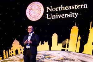 President Aoun standing on stage in front of a screen that shows a picture of the globe rising from the bottom of the screen with various silhouettes of famous world structures on it in yellow.