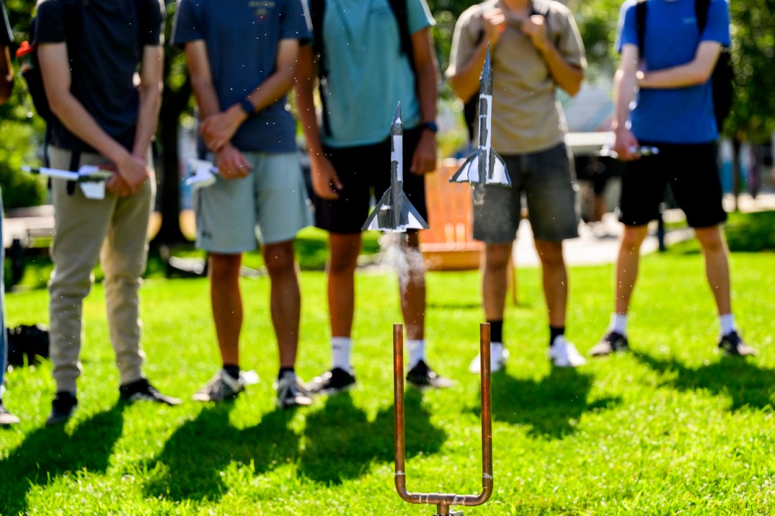 Students stand outside watching two model rockets blasting off.