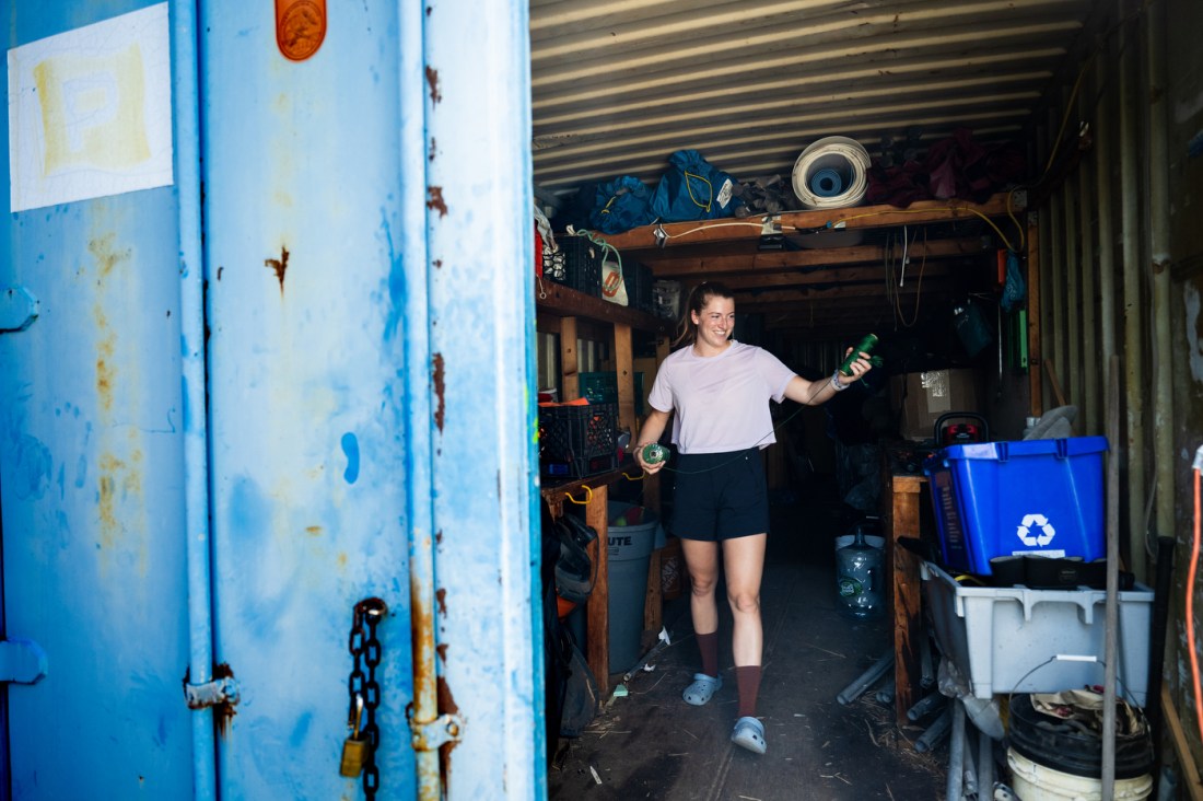 A woman stands inside a repurposed shipping container filled with tools and equipment. She appears to be organizing materials related to environmental work. The container is partially open, revealing recycling bins, coiled hoses, and supplies. The surrounding area looks industrial and functional, suggesting a fieldwork setting. Text over the image describes a coastal restoration project in Boston involving students and alumni from Northeastern University.