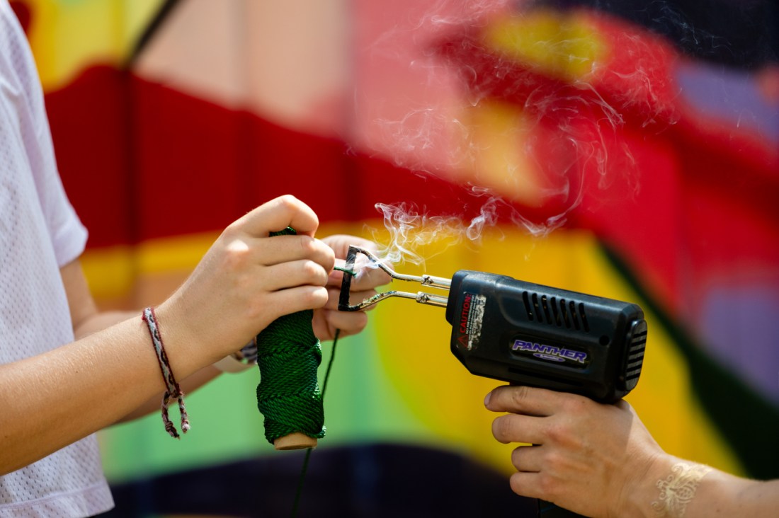 Two people hold a green, net-like rope structure outdoors in front of a vivid, colorful background. The image represents a project by Anja Clark, Kayla Sklar, and Gabriel Cira focused on restoring Boston’s coastline through nature-based infrastructure.