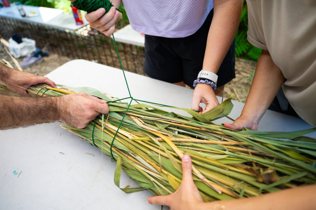 Three individuals work closely together weaving green reeds or long grass stems by hand. None of them are wearing gloves. Their hands are visible as they tie and bundle the materials with green string on a table. The project appears to be environmentally focused.