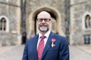 Portrait of Ken Henderson wearing a blue blazer and red tie standing outside a historic red building. On his lapel is a gold medal.